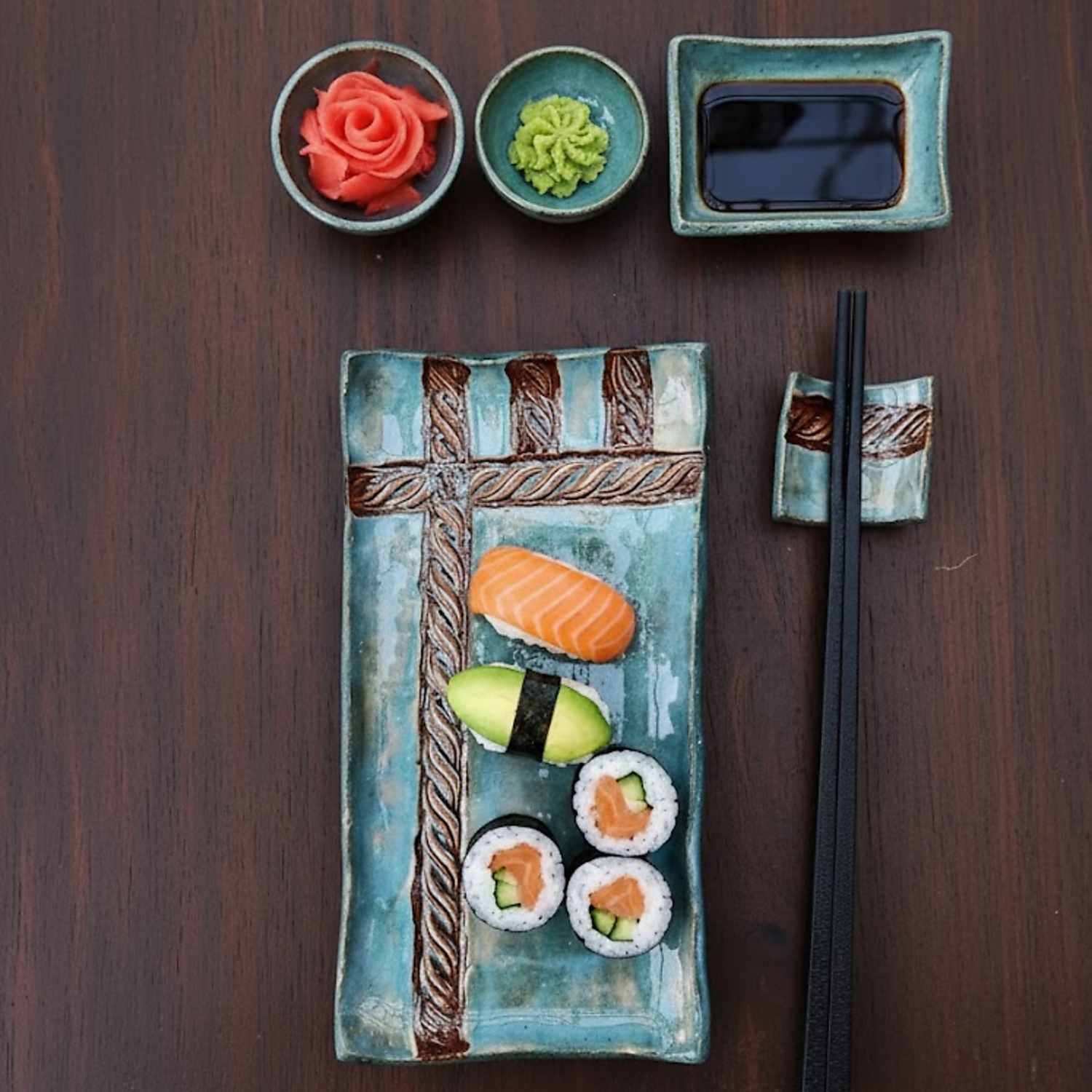 Handmade turquoise ceramic sushi set featuring a rectangular plate with an embossed rope pattern, wasabi and ginger bowls, and a soy sauce dish on a dark wood background.
