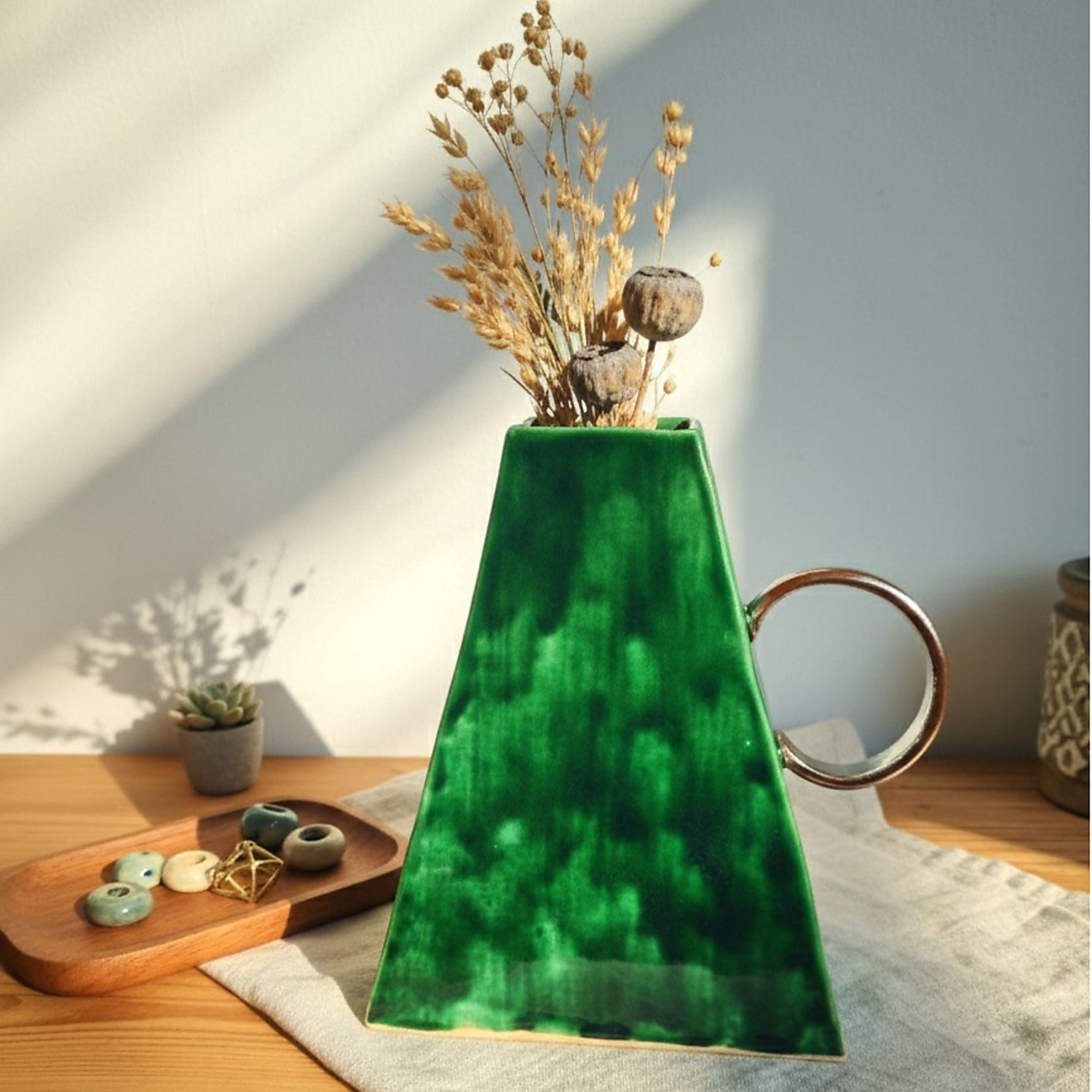 Handmade emerald green ceramic vase with a unique geometric triangle shape and a circular handle, styled with dried flowers on a wooden table.