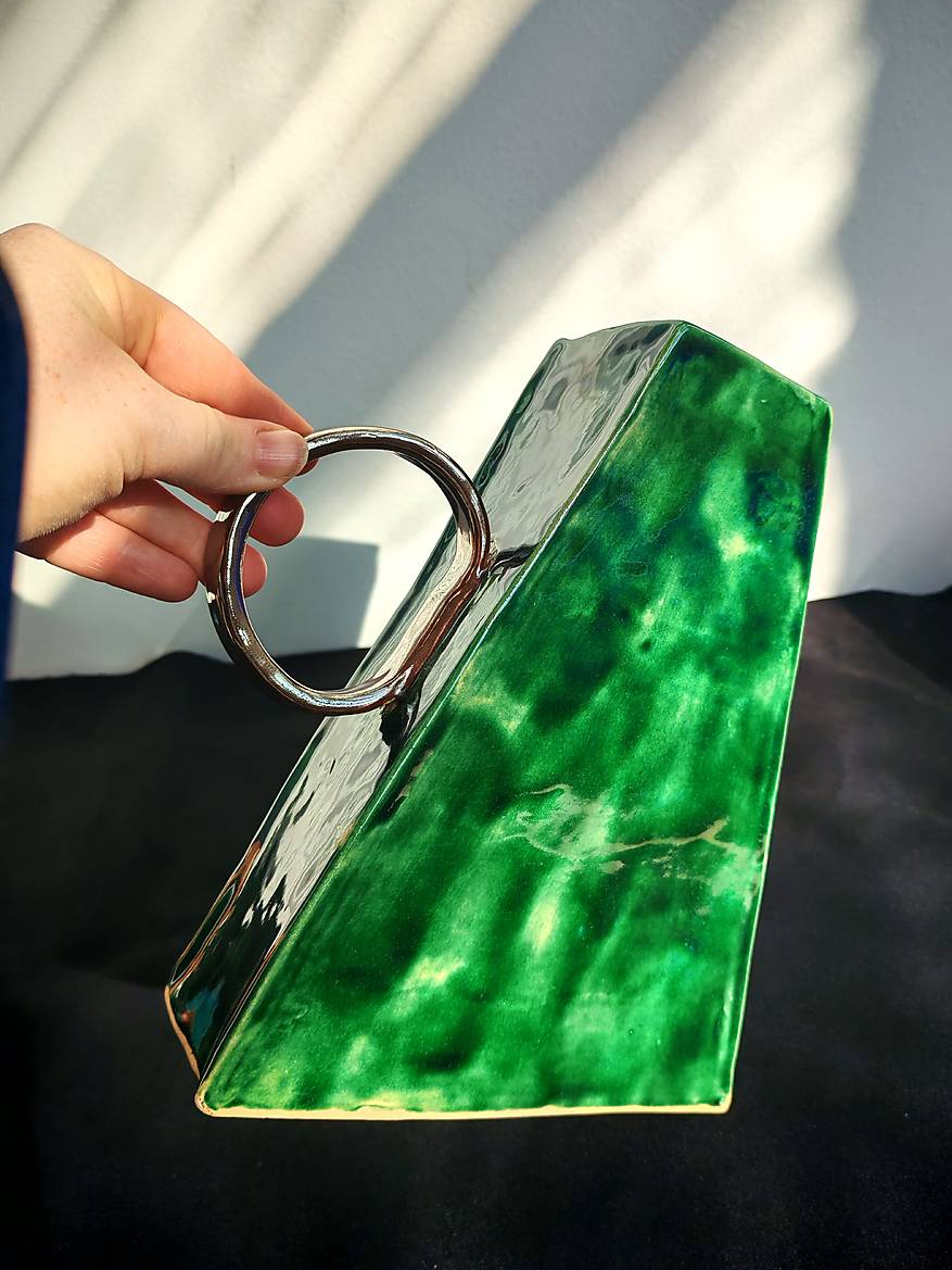 Green ceramic vase with a metallic handle held by a person against a neutral background