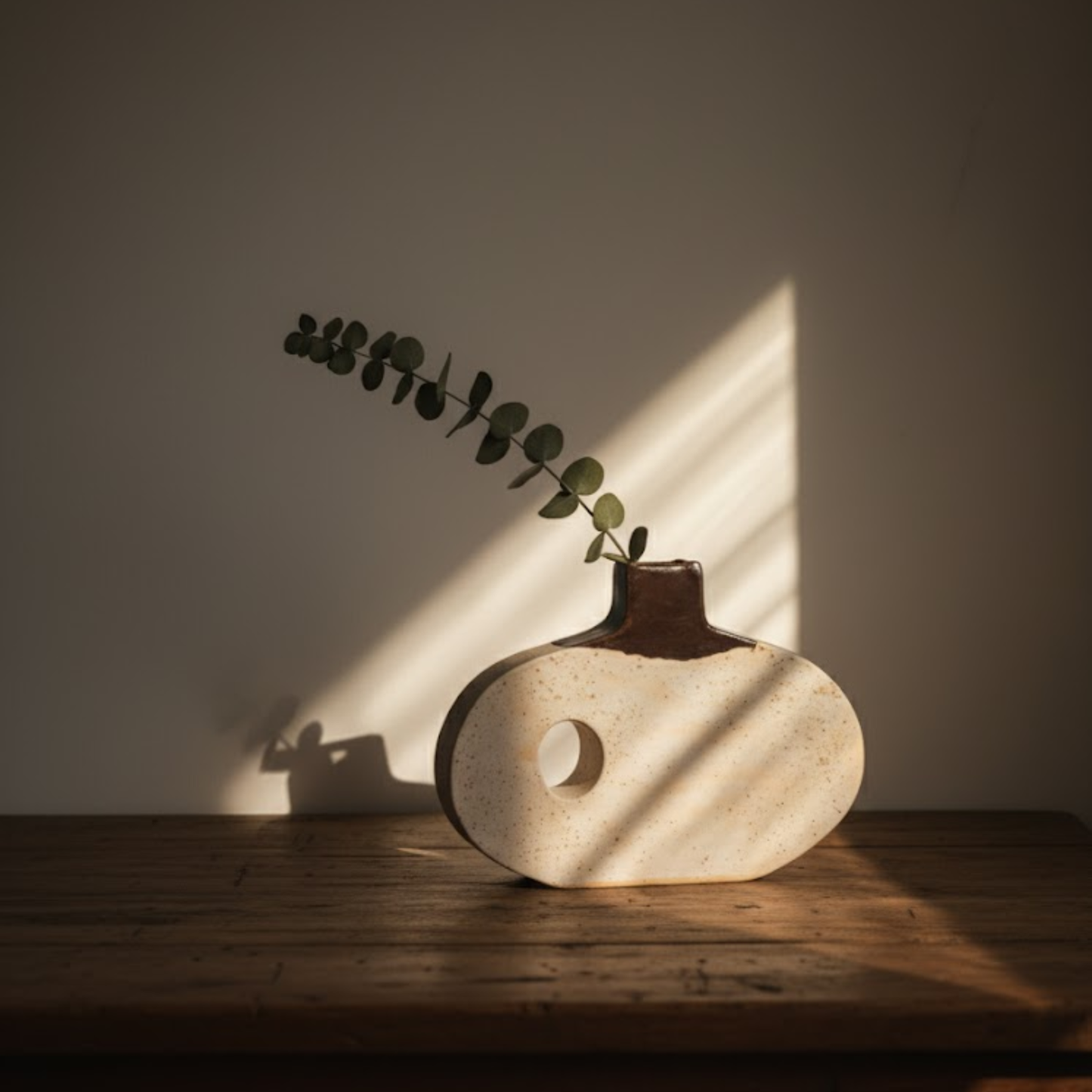 Minimalist handmade oval ceramic vase in speckled white with a dark brown neck and a circular cutout, styled with a eucalyptus branch on a wooden surface.
