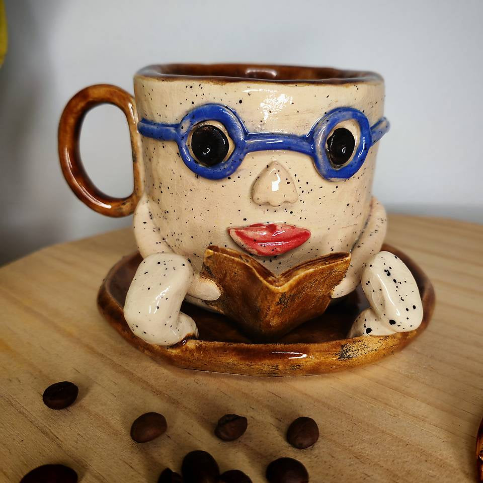 Handmade ceramic face mug featuring a 3D sculpted lady with blue glasses and red lips reading a book, on a rustic wooden background.
