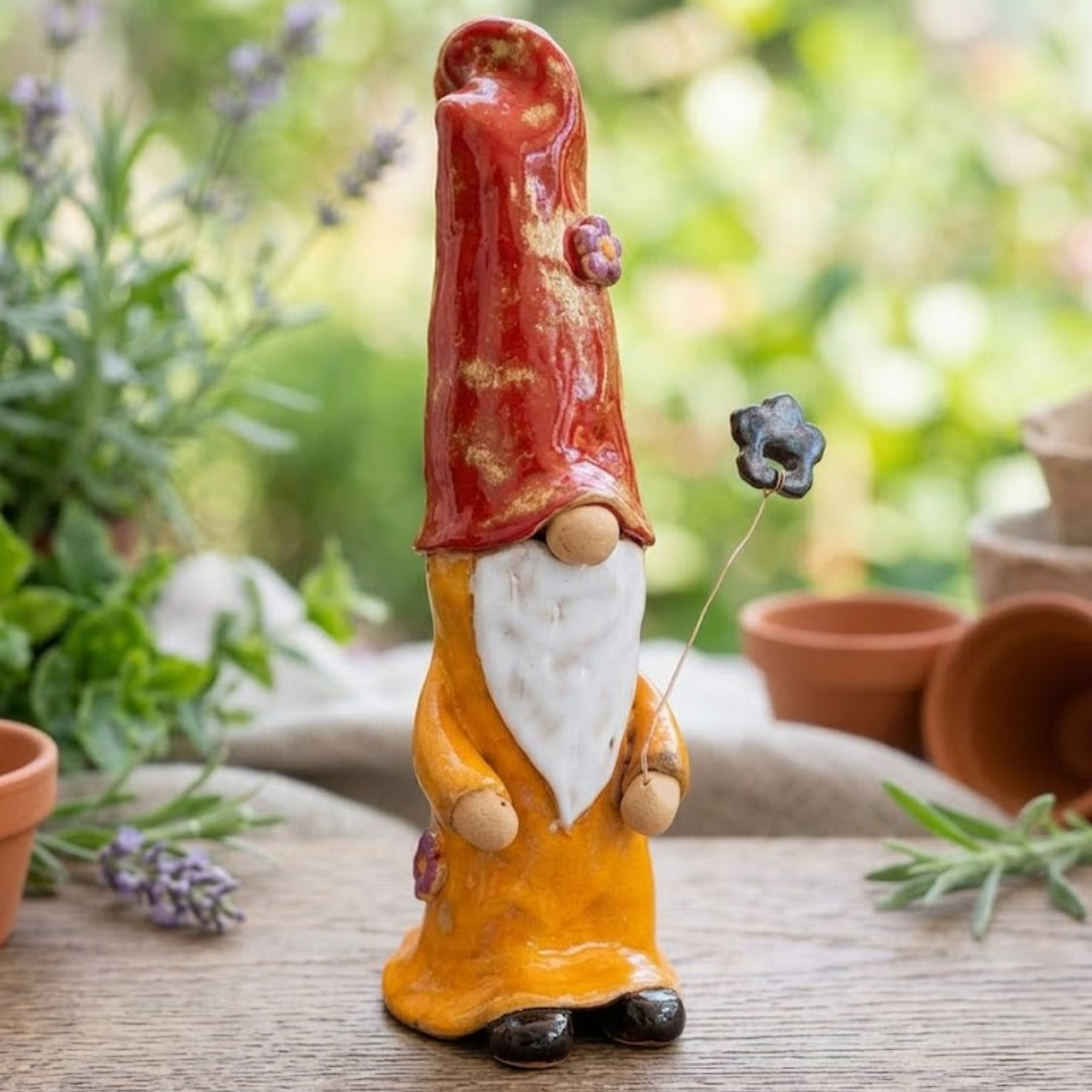 Handmade ceramic garden gnome with a tall red hat and yellow robe, holding a kinetic wire-mounted flower that moves in the wind, artisanal garden decor.